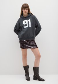 Felpa grigia con cappuccio e stampa bianca "91", abbinata a una gonna corta con paillettes in varie tonalità di marrone. Stivali neri al ginocchio.