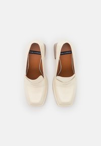 Une paire de mocassins à talons carrés en cuir de couleur crème, avec des orteils carrés, des coutures visibles et des semelles intérieures marron étiquetées "Ángel Alarcón".