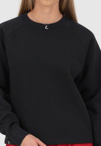 Felpa nera con scollatura a girocollo e maniche raglan. Tessuto morbido con polsini e orlo a coste. Indossata sopra un outfit rosso. Collana con pendente in argento.