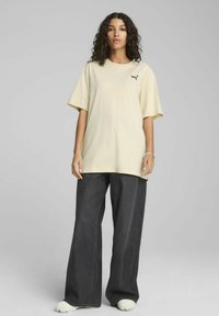 T-shirt oversize beige in tessuto morbido, con un piccolo logo sul petto, abbinato a pantaloni in denim scuri a gamba larga e sneaker chiare.