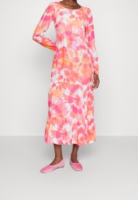 Robe longue à manches longues tie-dye dans des nuances vives de rose, orange et blanc, avec un ourlet à volants et une texture de tissu douce. Associée à des ballerines roses.