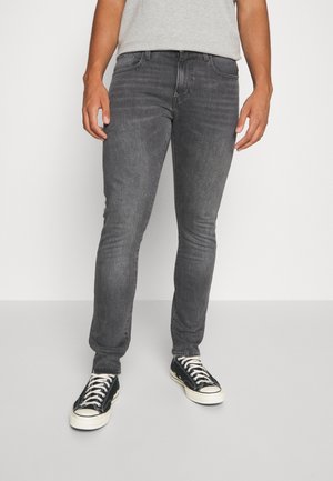 Persoon die nauwsluitende donkergrijze spijkerbroek draagt, een lichtgrijs shirt en zwarte lage sneakers met witte veters en zolen, staand op een lichte vloer.