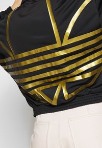 Black jacket with a glossy gold geometric pattern. Features elastic hem and cuffs, made from a smooth, lightweight fabric.