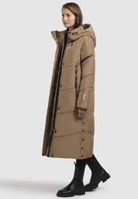 Garšs bežs pūkains mētājs ar kapuci, kas ir ar diagonālu šuvēm, melniem rāvējslēdzējiem, spiedpogu aizdari un sānu kabatām.