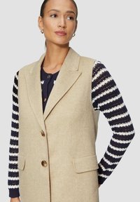 Beige Wollweste mit Schalkragen, zwei vorderen Taschen und einer einzelnen Knopfschließe, getragen über einem navy- und weißgestreiften Strickpullover.