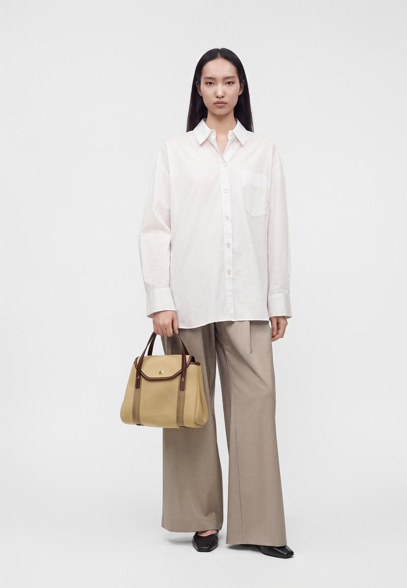 Femme aux longs cheveux noirs portant une chemise blanche oversize et un pantalon large beige, tenant un sac à main beige avec des anses marron.