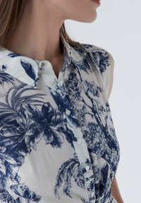 Chemise à manches courtes en tissu léger, arborant un motif floral bleu sur un fond clair, avec un col et sans manches.
