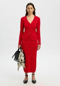 Gilet en tricot côtelé rouge avec une jupe midi assortie, associé à des ballerines noires et à un sac à main noir avec un détail en foulard à pois.