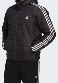 Zwarte gewatteerde jas met capuchon, met witte horizontale strepen op de mouwen en een Adidas-logo op de borst. Getextureerde zakken met rits.