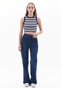 Maglietta a righe navy e bianca accostata a jeans flared in denim blu a vita alta e sneakers bianche. La modella è in piedi contro uno sfondo bianco.
