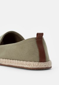 Olivgrön canvas slip-on sko med en beige rep-sula och brunt läderackent på hälen. Slät textur med minimalistisk design.