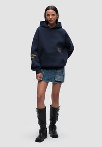 Felpa oversize blu navy in misto cotone, con tasca frontale e ricamo giallo. Abbinata a una gonna di jeans e stivali neri fino al ginocchio.