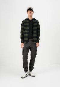 Zwart en groen geruit overhemd over een zwart T-shirt, grijze cargobroek met zijzakken, en witte sneakers met zwarte en grijze accenten.