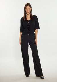 Cardigan nero lavorato a maglia con scollatura rotonda e maniche corte, dettagli con bottoni, abbinato a pantaloni neri a vita alta e gamba larga, e tacchi neri.