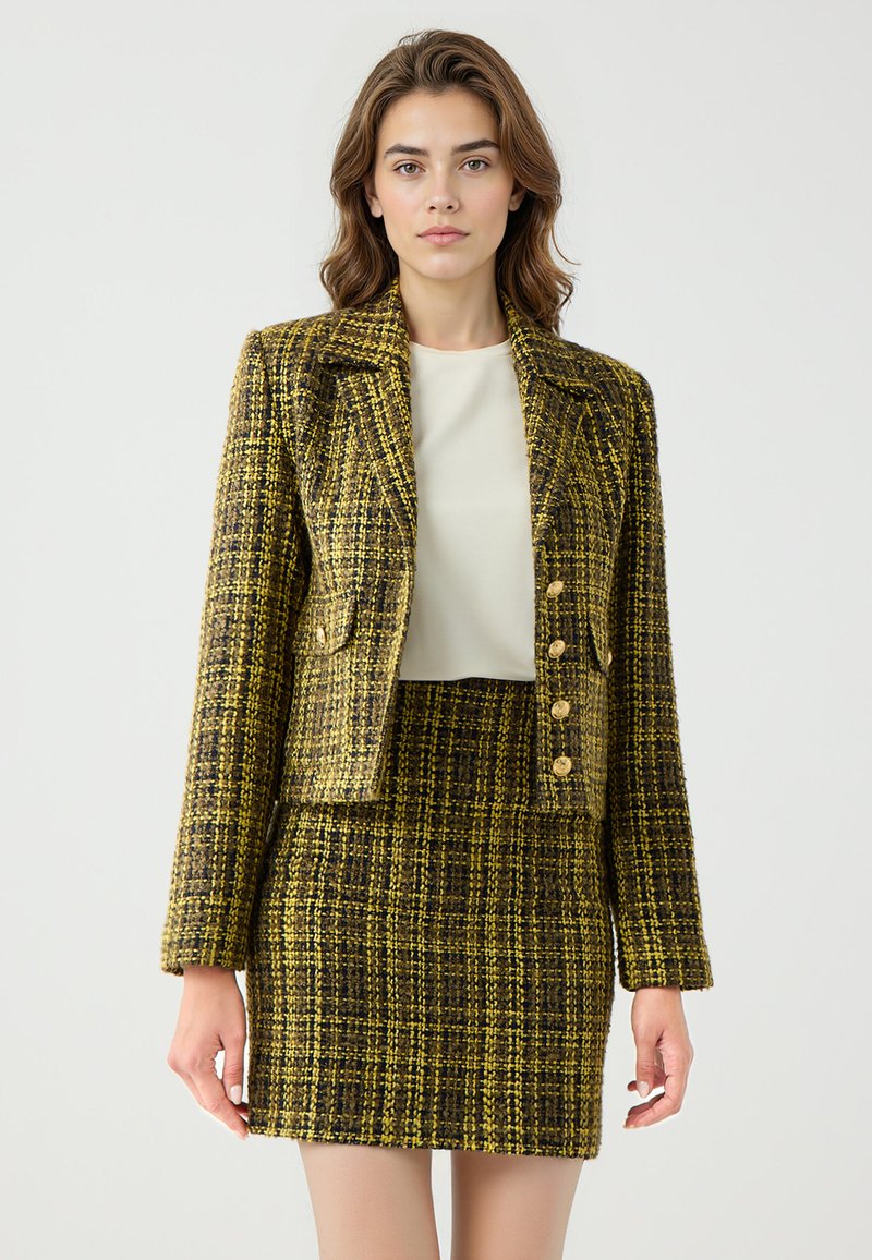 Wollen mix twee-delige pak in een geel-zwart ruitpatroon. Gestyled met een getailleerde blazer, opvallende knopen aan de voorkant en een bijpassende hoge taille rok.