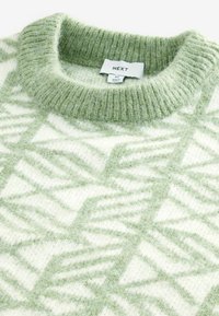 Pull en maille vert clair et crème, présentant un motif géométrique. Col côtelé et matière en laine douce et texturée. Étiquette de la marque visible.