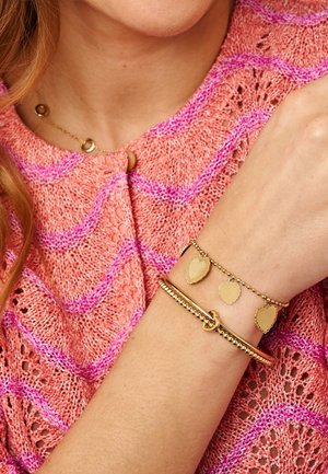 Femme portant un haut en tricot rose et violet avec des bracelets en or, dont un bracelet avec un pendentif en forme de cœur, à son poignet près de son cou.