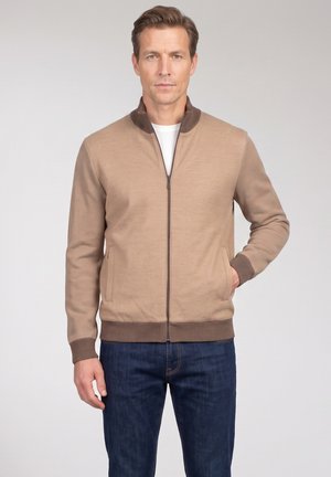 Mann trägt eine beige Jacke mit Reißverschluss, dunkelbraunen Bündchen und Kragen, kombiniert mit dunkelblauen Jeans, stehend vor einem schlichten Hintergrund.