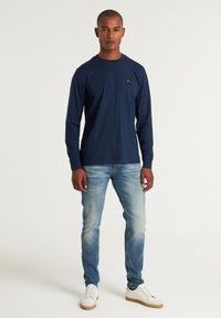Marineblaues Langarmshirt mit Rundhalsausschnitt, aus weichem Stoff und mit kleinem Logodetail. Kombiniert mit hellblauen Slim-Fit-Jeans und weißen Sneakers.