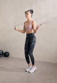 Atleta vestindo um top curto rosa e leggings preto camuflado, pulando corda. Ténis de treino cinzentos com detalhes brancos. Piso liso, fundo mínimo.