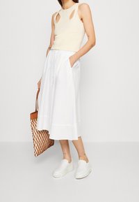 Top sin mangas beige con detalles recortados, falda midi blanca abotonada, zapatos blancos sin cordones y bolso tote tejido marrón.