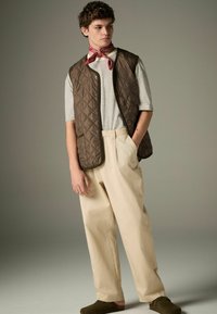 Quilted brown vest over a striped short-sleeve shirt, paired with beige trousers and dark green slip-ons, featuring a red bandana at the neck.