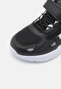 Basket noire avec tige en mesh et cuir synthétique, fermeture à lacets et Velcro, semelle blanche et accents texturés sur le bout et les côtés.