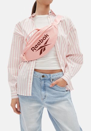 Frau trägt hellblaue Jeans, weißes bauchfreies Top, rosa-weiß gestreiftes Hemd und eine rosa Reebok Umhängetasche quer über der Brust.