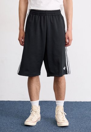 Mand iført sorte Adidas-shorts med hvide striber, hvide sokker og beige høje sneakers, stående på blå tæppe.