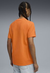 Mand med fletninger iført en orange kortærmet skjorte og lyseblå jeans, vendt væk fra en ensfarvet grå baggrund.