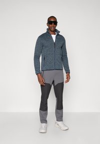 Fleece jacket in dark blue with a zip front and two side pockets; paired with gray and black color-blocked pants and white sneakers.