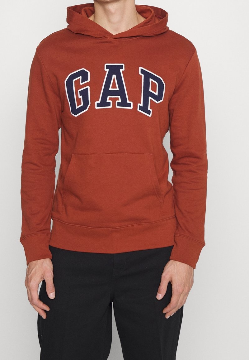 Sudadera de color naranja burno, material de mezcla de algodón, que presenta un bolsillo frontale y un gran logo de "GAP" en azul marino con contorno blanco. Capucha ajustable.