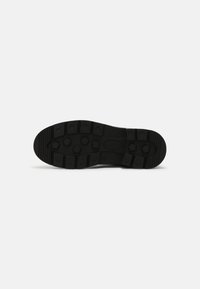 Semelle de chaussure en caoutchouc noir avec des crampons profonds présentant des motifs hexagonaux et rectangulaires sur fond blanc.