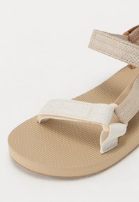 Beige Sandale mit strukturierten Gummisohle, metallischem Riemen und zwei verstellbaren Stoffriemen in hellbeige und braun.