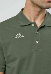 Mann som har på seg olivengrønn poloshirt med tre knapper og lite hvitt brodert logo av to sittende figurer på brystet.