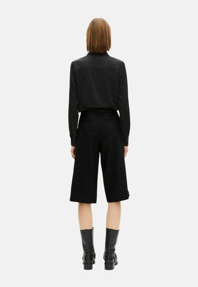 Camicia nera a maniche lunghe abbinata a pantaloni corti neri a gamba larga. Il tessuto è morbido e l'outfit presenta un design pulito. Stivaletti neri alla caviglia completano il look.