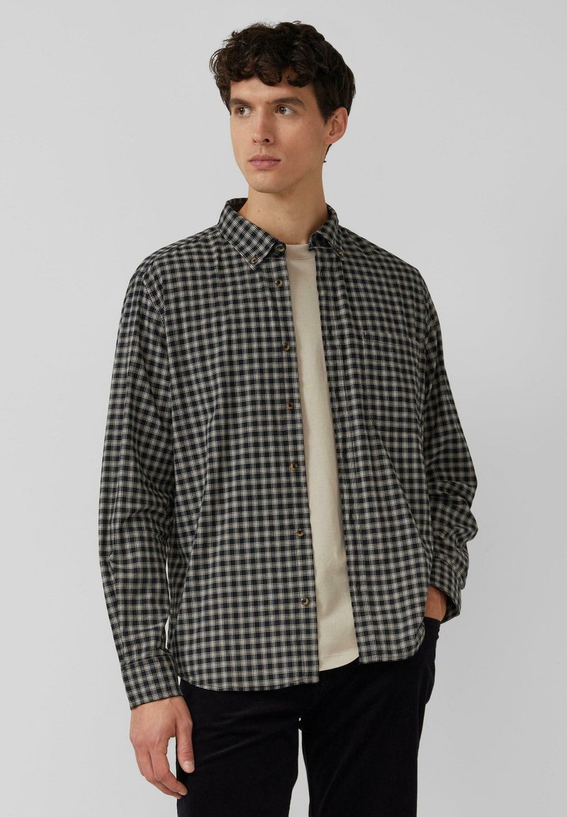 Schwarz-weißer karierthemd mit Button-Down-Kragen, langen Ärmeln und lockerer Passform, über einem hellbeigen T-Shirt getragen.