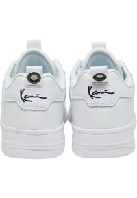 White sneakers with smooth synthetic material, padded collar, pull tab, black logo embroidery, and rubber sole.