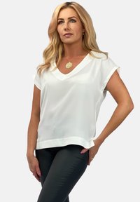 Biała, krótko rękawowa bluzka z dekoltem w serek, o miękkiej fakturze i luźnym kroju; w połączeniu z dopasowanymi ciemnymi spodniami.