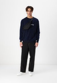 Marineblå sweater med struktureret mønster, rund hals og logo detalje. Sort crossbody taske båret diagonalt. Sorte bukser og hvide sneakers.