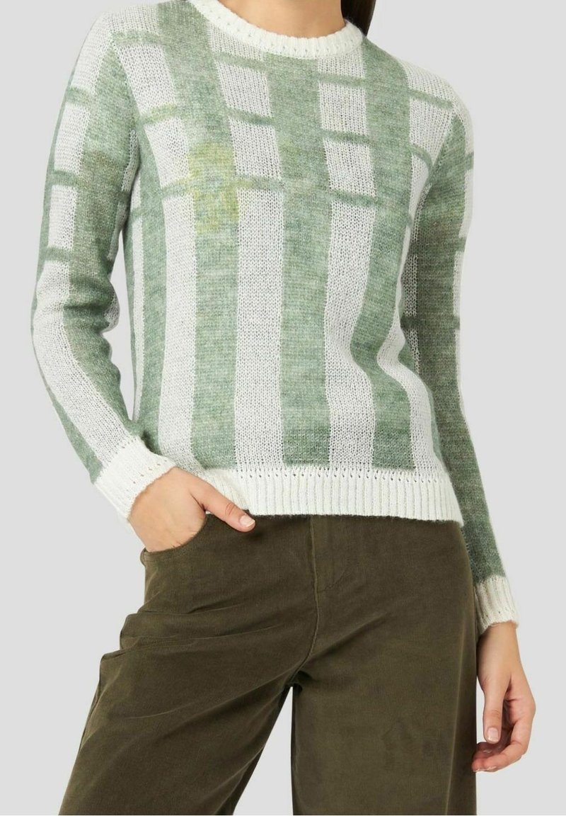 Femme portant un pull en maille rayé vert et blanc et un pantalon vert olive, avec une main dans la poche, sur un fond uni.