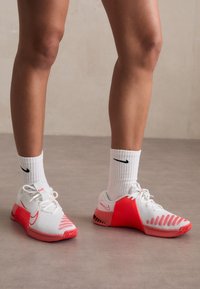 Zapatillas deportivas blancas con detalles en rosa, parte superior de malla texturizada, suela de goma y cordones. Llevadas con calcetines blancos acanalados con un logo negro.