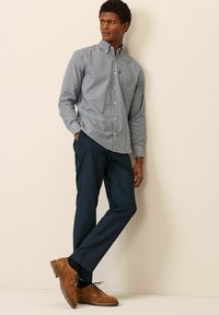 Chemise à carreaux blanche et bleue, pantalon navy, chaussures en daim marron, appuyé contre un mur, les mains dans les poches.