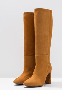 Botas altas de ante para mujer en marrón claro con punteras puntiagudas y tacones altos de bloque, colocadas una al lado de la otra sobre una superficie blanca.