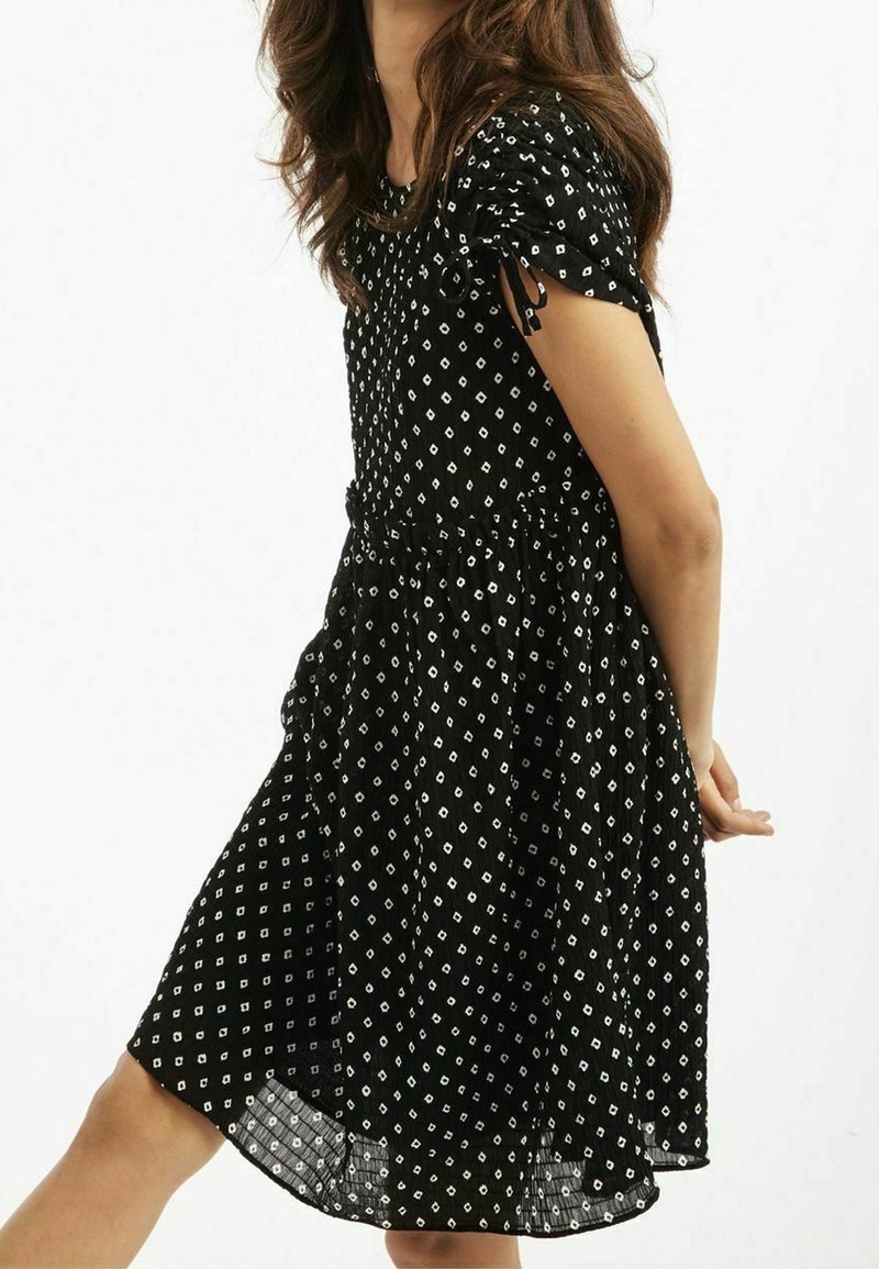 Robe noire avec un corsage ajusté, des manches courtes bouffantes et une jupe évasée. Présente des motifs en forme de diamants blancs et une taille froncée.