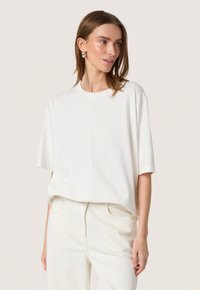 Hvid bomulds t-shirt med korte ærmer, afslappet pasform, rund halsudskæring og diskret logo detaljer på ærmet. Parret med lyse bukser.