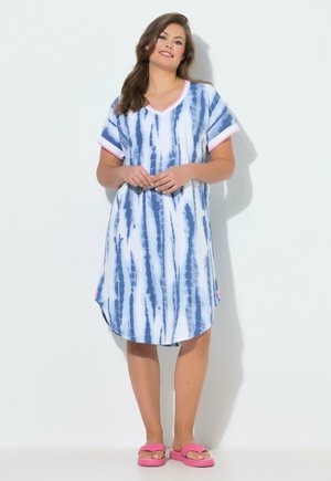 Kobieta w niebiesko-białej sukience do kolan z efektem tie-dye z różowymi wykończeniami i różowymi klapkami, stojąca na białym tle.