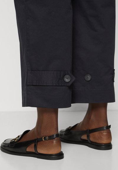 Spodnji nogi oblečeni v črne skrajšane hlače z gumbi in črne usnjene slingback loafer čevlje z zlatimi sponkami.