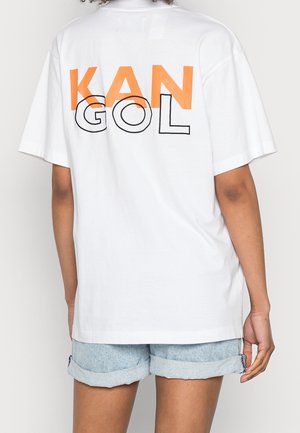 Personne portant un t-shirt blanc oversize avec "KANGOL" en lettres orange et noires dans le dos, associé à un short en jean clair retroussé.