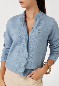 Vrouw in een lichtblauwe gebreide cardigan met kantpatroon en golfrandjes, beige broek en een chunky gouden armband om haar pols.